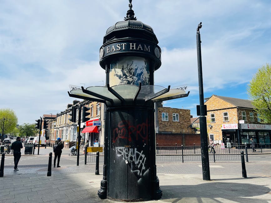A cylindrical street clock with a domed top, displaying the words East Ham, is situated on a paved sidewalk in a busy urban area. The clock is mounted on a black metal stand, which features a mesh panel with graffiti tags. Surrounding the clock are bollards and metal fencing, separating the pedestrian pathway from the street. In the background, there are several pedestrians walking, some standing near the crosswalk, and a row of shops including a nail salon on the right. The sky above is partly cloudy with patches of blue visible. The scene depicts typical city street activity, with buildings made of brick and a mix of commercial and residential properties. This image, emphasizing urban infrastructure and pedestrian movement, supports content related to city living and relocation services, as offered by Man and Van Bromley.