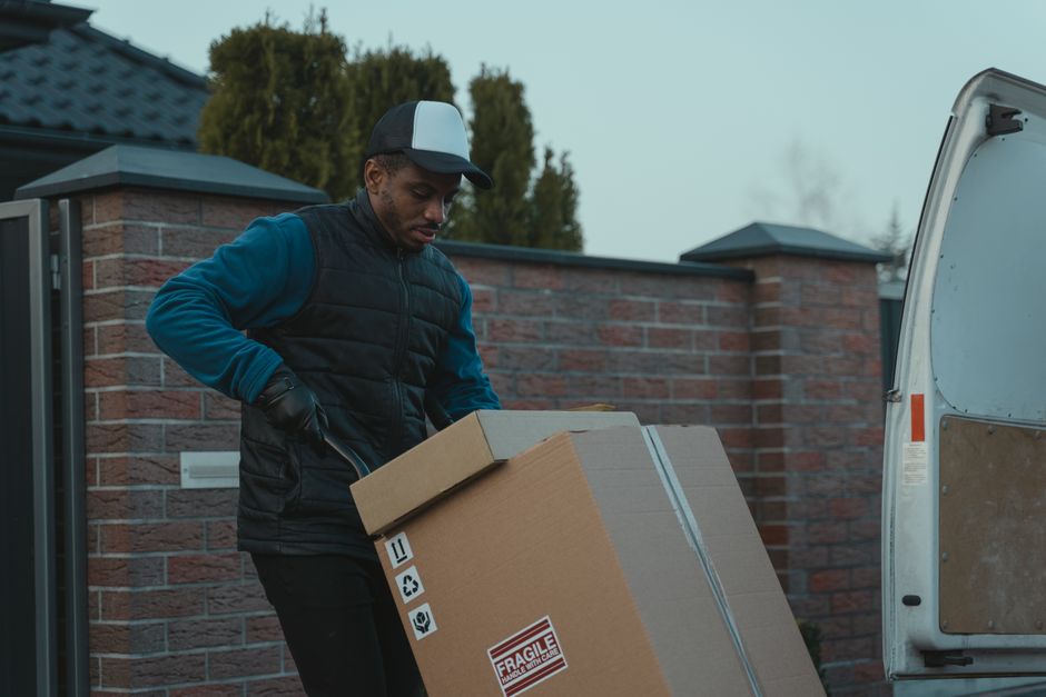 A man wearing a black quilted vest, blue long-sleeve shirt, black gloves, and a baseball cap is standing outdoors at a residential property, loading a large cardboard box wrapped with grey packing tape onto a van. The box features various handling symbols, including a fragile warning, recycling icon, and a symbol indicating careful handling. The background shows a brick wall with a dark-colored gate and a roof, with trees in the distance. The scene is set during the day under diffuse natural light, illustrating a packing and loading process involved in house removals or furniture transport, with the man from Man and Van Bromley preparing items for a home relocation.
