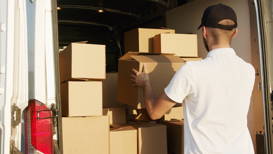 A man wearing a black cap and a white t-shirt is inside the back of a van, which is parked near a building with a doorway visible. He is holding a large, light brown cardboard box, carefully positioning it among other similar boxes within the vehicle. Several stacked cardboard boxes, some with open flaps, are arranged in the van, ready for loading or unloading. The interior of the van is dark, with some scattered packing materials and possibly protective blankets or bubble wrap visible around the boxes, indicating a home relocation or furniture transport process. The scene is illuminated by natural daylight, highlighting the details of the boxes, the man's clothing, and the vehicle interior. This image is associated with professional removals services, such as those offered by Man and Van Bromley, for house moves and small load relocations. It captures the careful handling and organized packing involved in moving logistics.